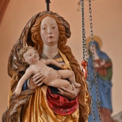 Eine Madonnafigur, die die Maria darstellt, mit langen, blonden, lockigen Haaren und einem braunen Tuch über den Haaren und einem gold-rotem Gewand, hat das nackte Jesusbaby mit gelocktem Haar im Arm. Die Figur ist an eine Halterung hängend befestigt. Daneben hängt eine Gitter-Kette. Im Hintergrund ist eine an die Wand gemalte Frauenfigur mit Heiligenschein im langen Gewand.