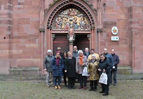 Der Vorstand und die Mitglieder des Kirchenbauverein Kiedrich e.V. stehen in Wintermänteln vor der Kirche mit roten Steinen, vor einem Holztor mit rotem Schmiedeschmuck und dem halbrundem Tympanon mit der Figur des Valentin und der Darstellung von mehreren Engeln, dem Jesukind und Gott.