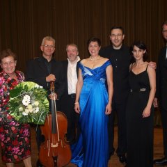 Vier Männer im dunklem Anzug und einem Cello und einer Frau im königsblauem Abendkleid und einer Frau im schwarzem Abendkleid sowie einer Dame im Kostüm mit rot- und pinken Blumen auf schwarzem Untergrund mit einem Blumenstrauß in der Hand mit weißen Blumen, stehen nebeneinander.