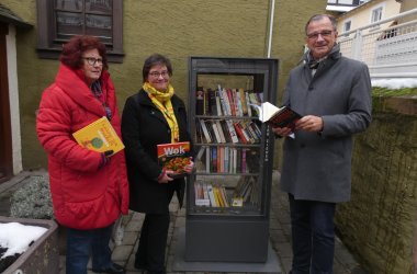 Bürgermeister Steinmacher steht vor einem mit Büchern gefüllten Bücherschrank aus Metall und mit Glastüren, neben zwei Frauen in Winterjacken mit Büchern in der Hand.    