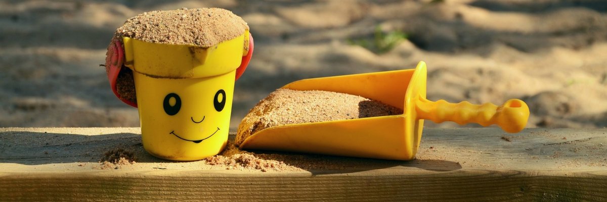 Auf einem Holzbalken, vor und hinter dem sich Sand befindet, liegt eine gelbe Sandspielzeugschaufel und ein gelber Sandspielzeugeimer mit einem Gesicht und einem roten Henkel. Die Schaufel und der Eimer sind mit Sand gefüllt.