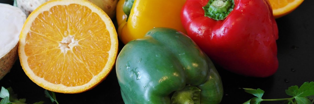 Eine rote, gelbe und grüne Paprika liegen bei einer halbierten Orange, geschälten Walnüssen, einem Knollensellerie und einem grünem Salat.
