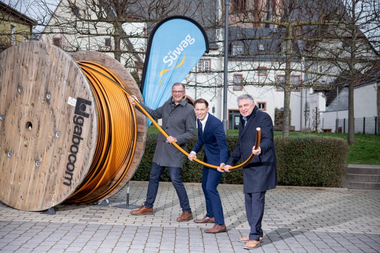 Herr Bürgermeister Steinmacher und zwei Mitarbeiter der Süwag Energie AG, gekleidet mit dunklen Anzugshosen und Mänteln oder Jackets stehen vor dem blauen SÜWAG Dropflag und ziehen an dem Ende eines orangenem Glasfaserkabels, das auf einer großen Holzspule aufgewickelt ist.  